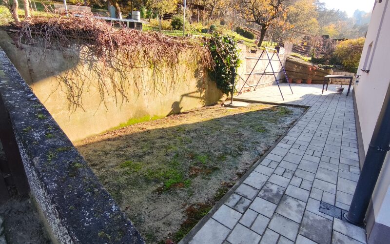 Úzky tienistý priestor pri dome s riedkou trávou a suchou pôdou – problémové miesto vhodné pre bezúdržbový trávnik